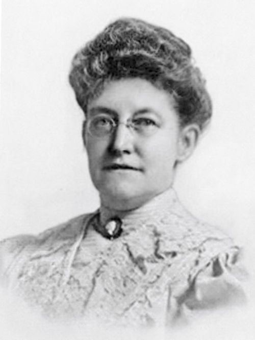 Black and white portrait of a woman with glasses and a high-collared dress, smiling softly at the camera.