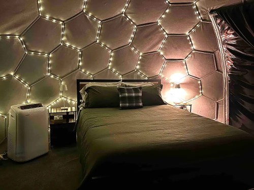 Cozy bedroom in a geodesic dome, featuring a bed, bedside tables, and warm string lights on the walls.