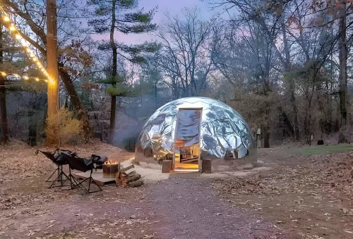 A geodesic dome in a wooded area, with smoke rising, chairs nearby, and string lights illuminating the scene at dusk.