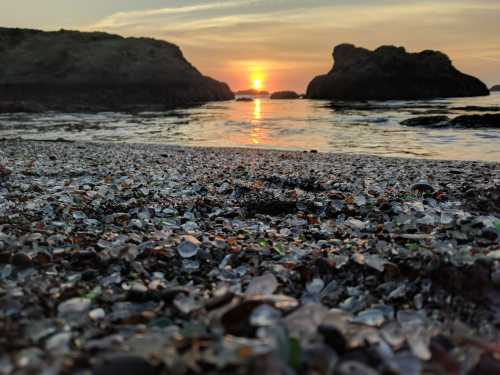 A beach covered in colorful glass shards, with a sunset reflecting on the water in the background.