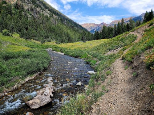A serene mountain landscape featuring a flowing river, lush greenery, and distant peaks under a blue sky.