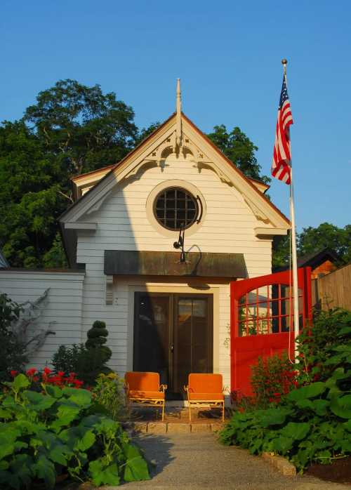 A charming white house with a circular window, red gate, and American flag, surrounded by greenery and flowers.