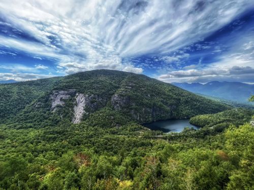 A panoramic view of a lush green mountain with a lake, under a dramatic sky filled with clouds.