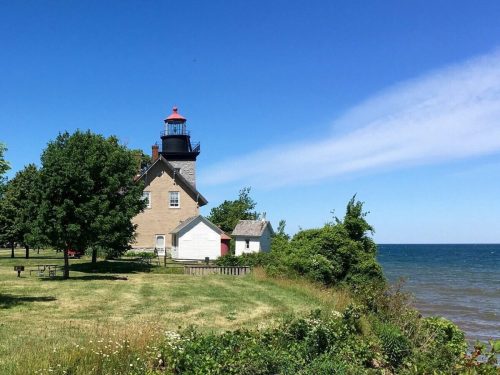 A lighthouse stands near the shore, surrounded by green trees and a clear blue sky over the calm water.