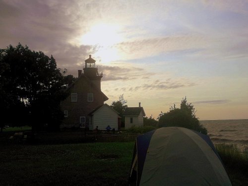 A lighthouse stands near the shore at sunset, with a tent set up in the foreground and calm waters in the background.