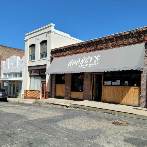 Exterior of Gooney's Bar & Grill with a gray awning, surrounded by historic buildings under a clear blue sky.