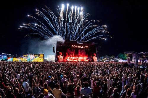 A large crowd at a music festival with fireworks lighting up the night sky above the stage.