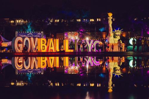 Colorful "GOVBALL NYC" sign illuminated at night, reflecting in water, with a statue resembling the Statue of Liberty nearby.