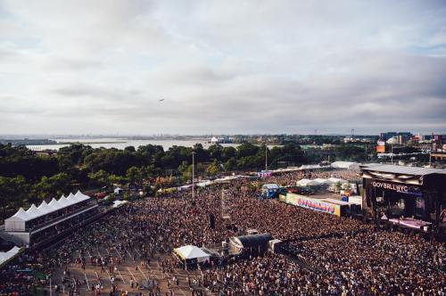 Aerial view of a large crowd at a music festival, with stages and tents set up against a cloudy sky.