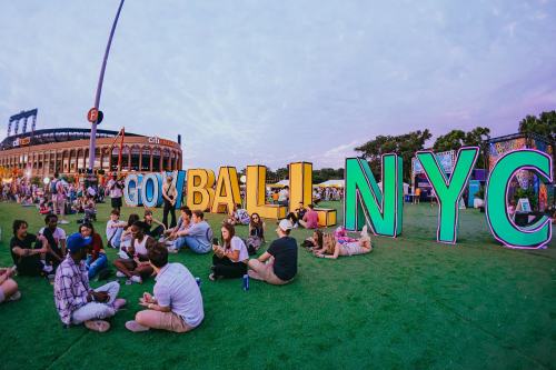 A vibrant outdoor event scene with people sitting on grass near large colorful letters spelling "GO BALL NYC."