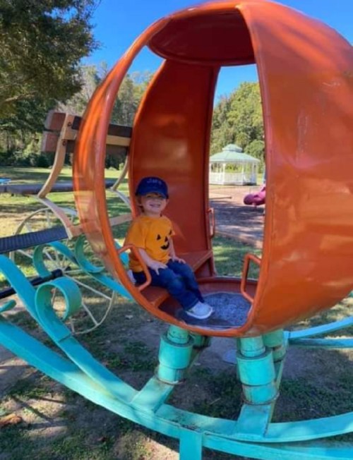 A child in a pumpkin-shaped play structure at a park, wearing a pumpkin shirt and a blue cap, smiling happily.