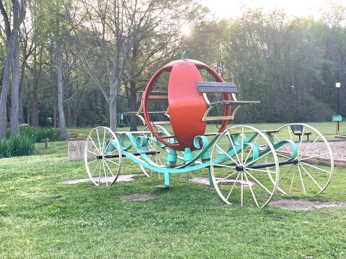 A whimsical pumpkin-shaped carriage on wheels, set in a grassy park surrounded by trees.