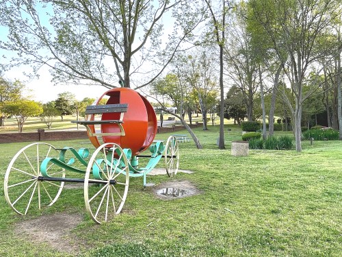 A colorful, oversized pumpkin carriage sits on a grassy area surrounded by trees in a park.