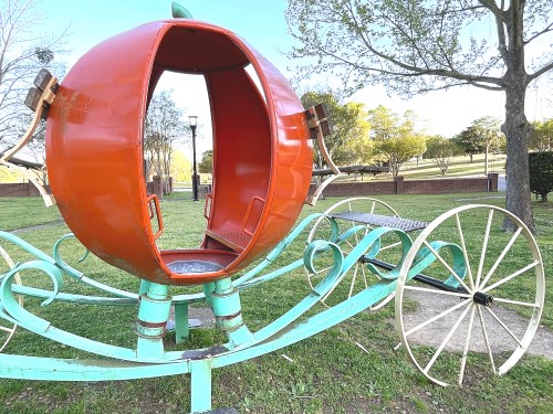 A whimsical pumpkin carriage structure in a park, featuring an open interior and decorative wheels.