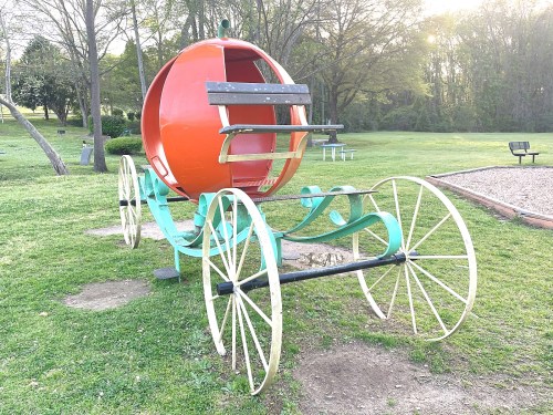 A whimsical orange pumpkin carriage on wheels, set in a grassy park with trees in the background.