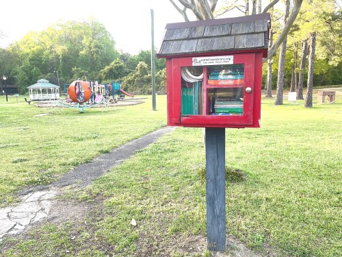 A small red library box filled with books, located in a park near playground equipment and trees.