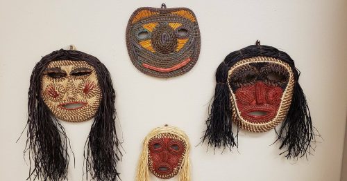 Four decorative masks made from natural materials, featuring various facial designs and colors, displayed on a wall.