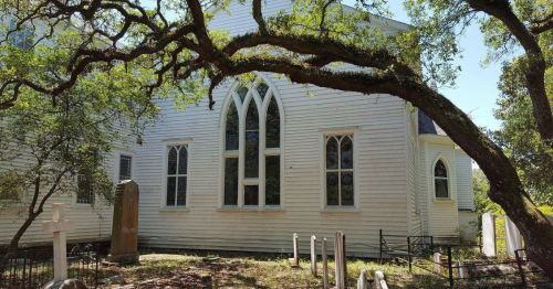 A white church with Gothic windows, surrounded by trees and gravestones in a peaceful setting.