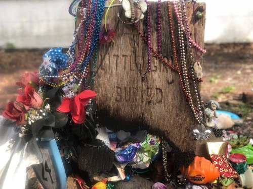 A weathered wooden grave marker surrounded by colorful beads, flowers, and various trinkets.