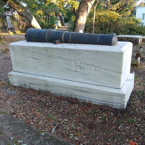 A marble grave marker with the name "Burns" and a cannon resting on top, surrounded by trees and a gravel path.