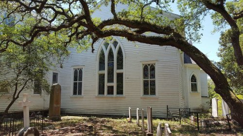 A white church with arched windows, surrounded by trees and gravestones in a peaceful outdoor setting.
