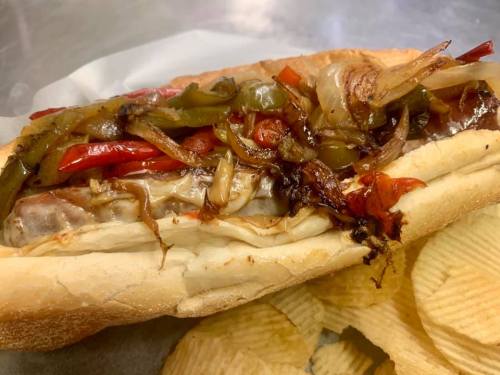 A loaded sandwich with grilled onions and peppers, served with a side of potato chips.