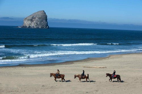 Three horseback riders stroll along a sandy beach with a large rock formation in the background and gentle ocean waves.