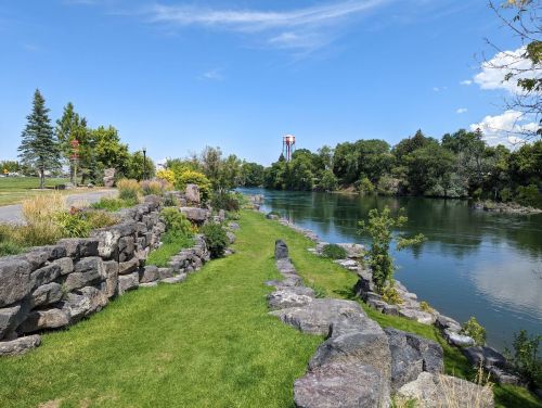A serene riverbank scene with lush greenery, stone pathways, and a clear blue sky in the background.