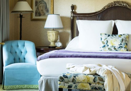 A cozy hotel room featuring a bed with white linens, a floral pillow, a blue chair, and elegant decor.