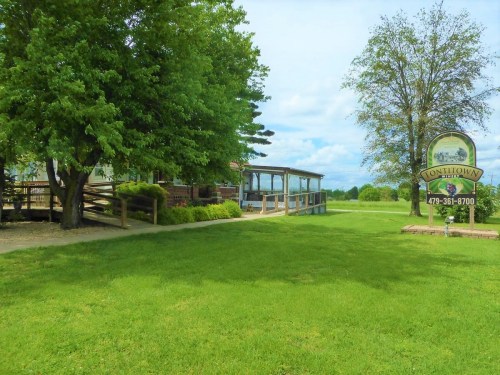A green lawn with trees, a building with a porch, and a sign for Tonitown under a blue sky with clouds.