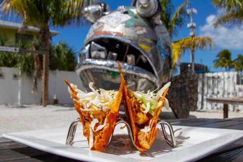 Two crispy tacos filled with fish and topped with shredded cabbage, set against a quirky metallic fish sculpture.