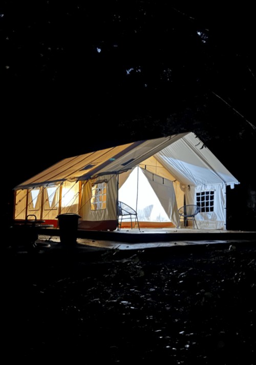 A lit tent at night, with glowing windows and chairs outside, surrounded by dark trees.