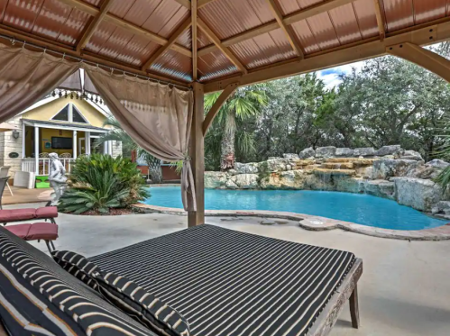 A shaded patio with a striped lounge chair overlooks a serene pool surrounded by rocks and lush greenery.