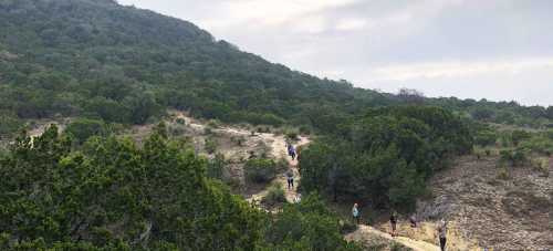 A winding trail through green hills with hikers walking along the path under a cloudy sky.