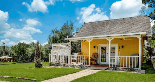 A charming yellow cottage with a porch, surrounded by green grass and trees under a bright blue sky with fluffy clouds.