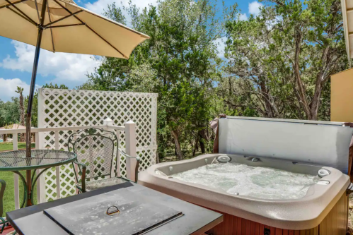 A hot tub on a deck surrounded by trees, with a table and umbrella nearby under a blue sky.