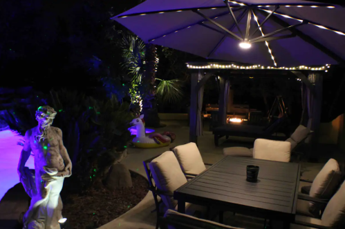 A cozy outdoor patio with a table, chairs, a statue, and string lights, surrounded by lush greenery and a fire pit.
