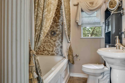 A cozy bathroom featuring a bathtub with a decorative curtain, a toilet, and a window with soft drapes.
