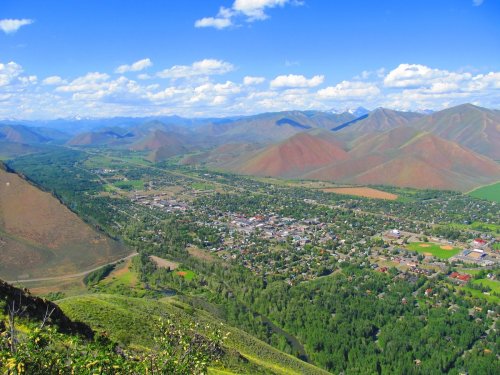 A panoramic view of a valley with a small town surrounded by green trees and rolling mountains under a blue sky.
