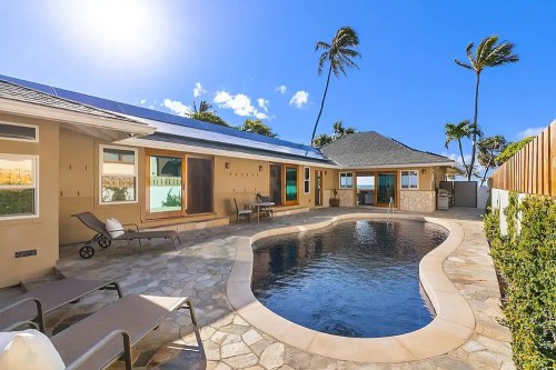 A sunny backyard with a stone patio, a swimming pool, and palm trees, featuring a house with large windows.