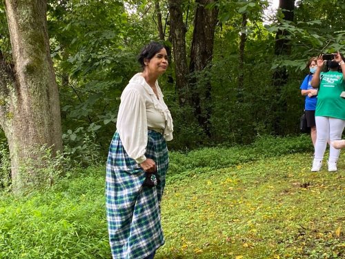 A woman in a historical outfit stands in a green, wooded area, with people observing in the background.