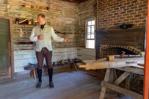 A man in historical attire stands in a rustic room with wooden walls, gesturing towards a workbench and fireplace.