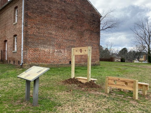 A wooden structure resembling a stocks, with a nearby informational sign, set against a brick building and cloudy sky.