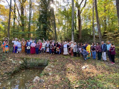 A large group of people in traditional attire gathered in a forested area, with autumn leaves and a small pond nearby.