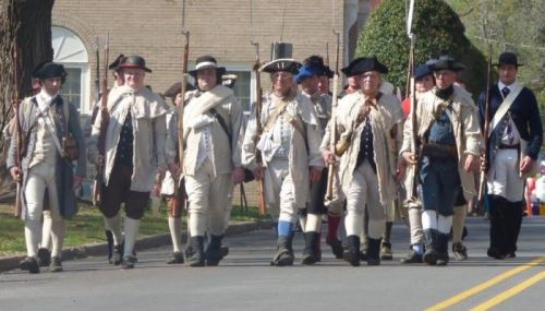 A group of men in historical military uniforms marching down a street during a reenactment event.