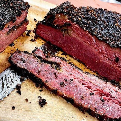 Close-up of sliced brisket with a smoky crust, resting on a wooden cutting board. Juicy and tender meat visible.