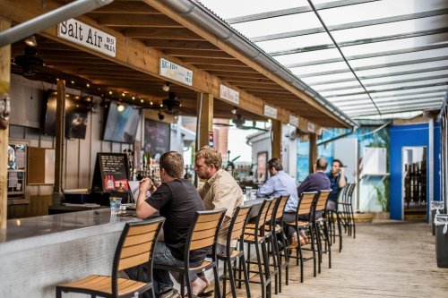 A lively bar with patrons seated at a long counter under a covered patio, enjoying drinks and conversation.