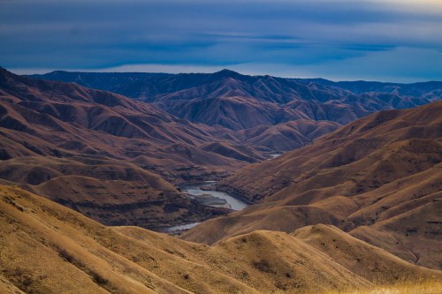 A panoramic view of rolling brown hills and a winding river under a blue sky, showcasing a serene mountainous landscape.
