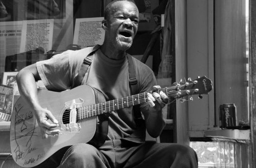 A man passionately sings and plays guitar on the street, with a can of soda nearby and historical photos in the background.