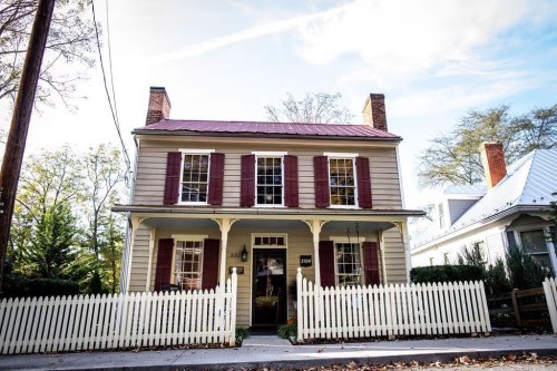 A charming two-story house with a red metal roof, white picket fence, and maroon shutters on a sunny day.